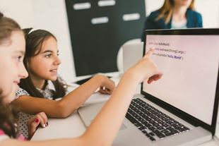 Young girls coding on laptop computer with teacher in the background