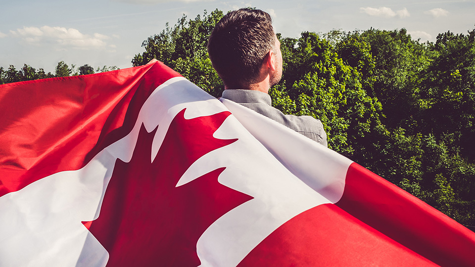 un homme tiens le drapeau du Canada sur son dos en regardant les arbres.