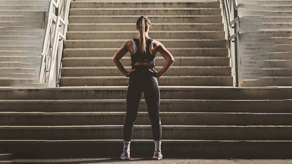 Une femme en vêtement de sports se tiens devant des escaliers avec ses mains sur ses hanches.